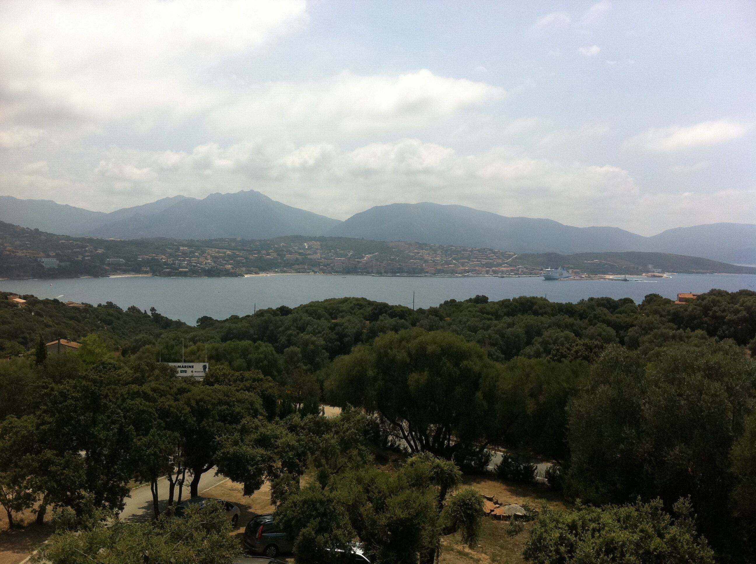 A coastal landscape with a bay, mountains in the background, and dense green trees in the foreground.