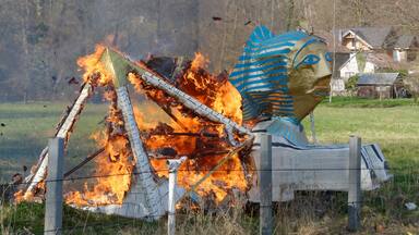 Ein großes, brennendes Abbild mit einem blau-weiß gestreiften Kopf, vor einer ländlichen Kulisse mit Häusern und einem Zaun.