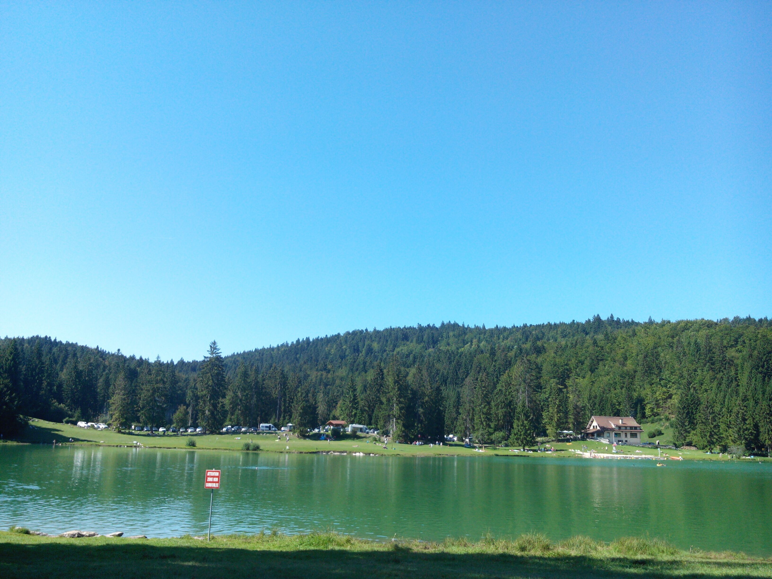 A lake surrounded by forested hills, a clear sky, and a few buildings in the distance.