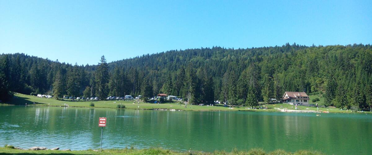 A lake surrounded by forested hills, a clear sky, and a few buildings in the distance.