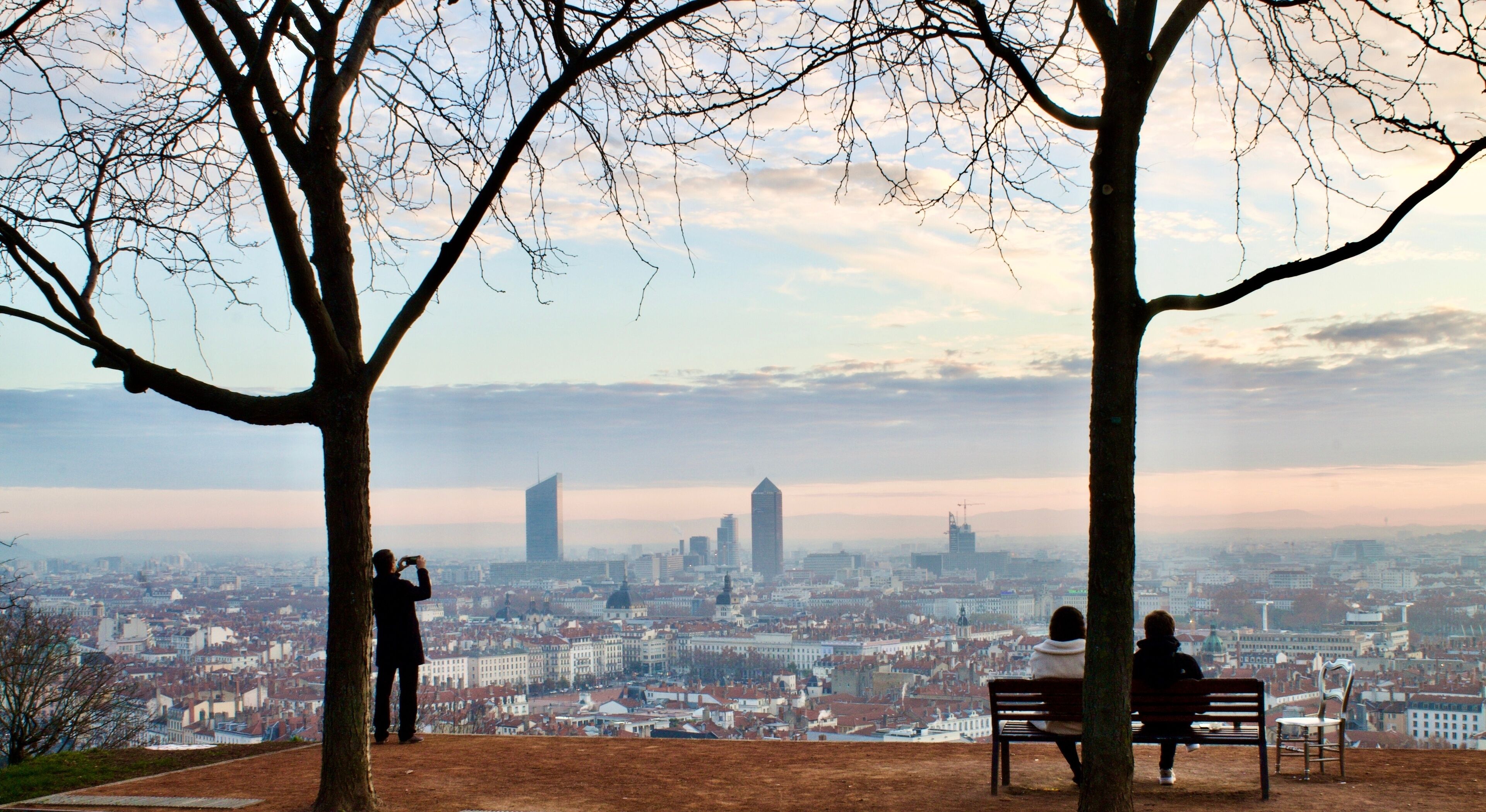2 people sitting on a bench admiring a wonderful sunrise over the city of Lyon in France with a tourist taking picture of the view
