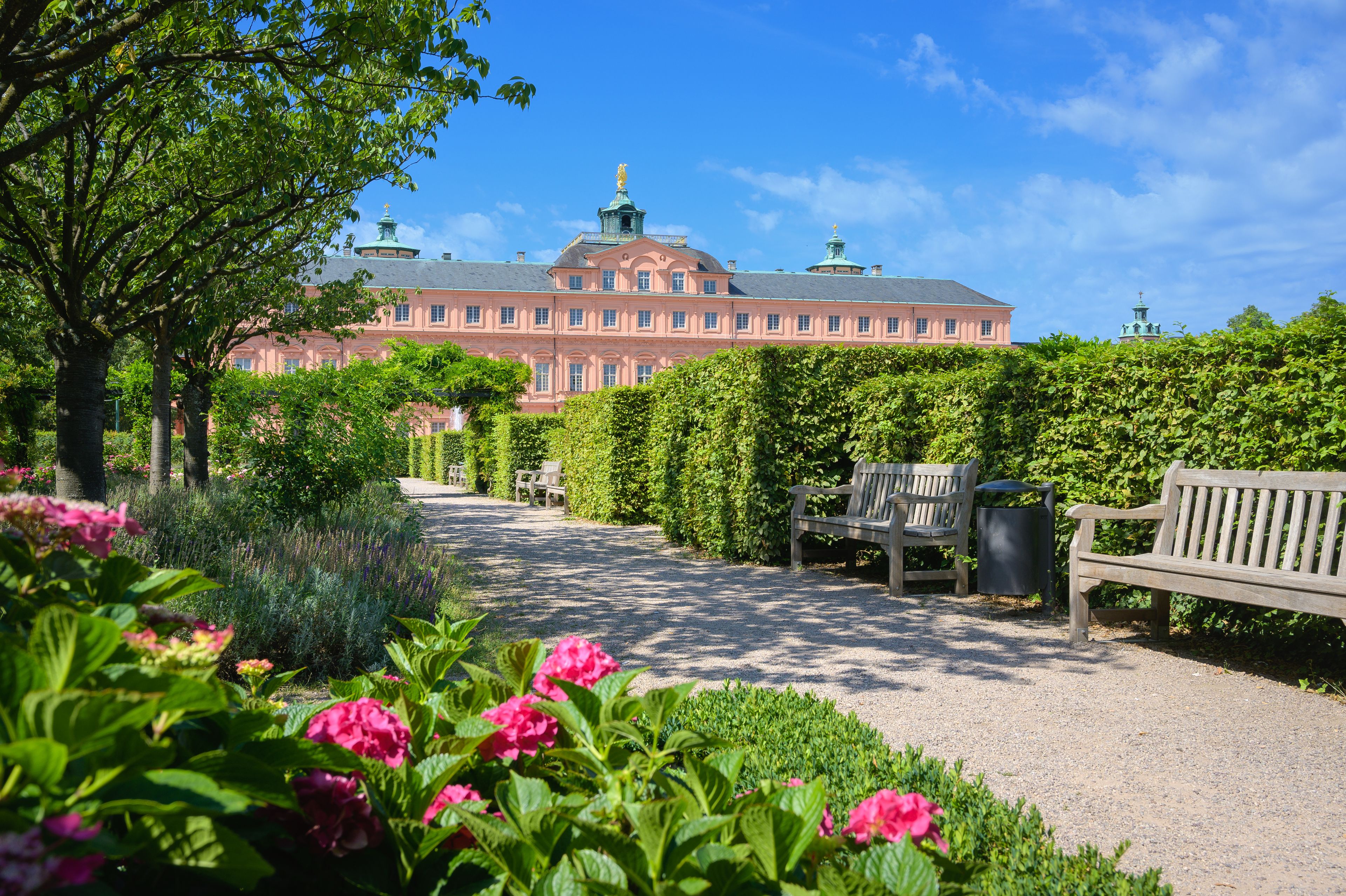 A large pink building with a green roof, surrounded by lush greenery and a gravel path.