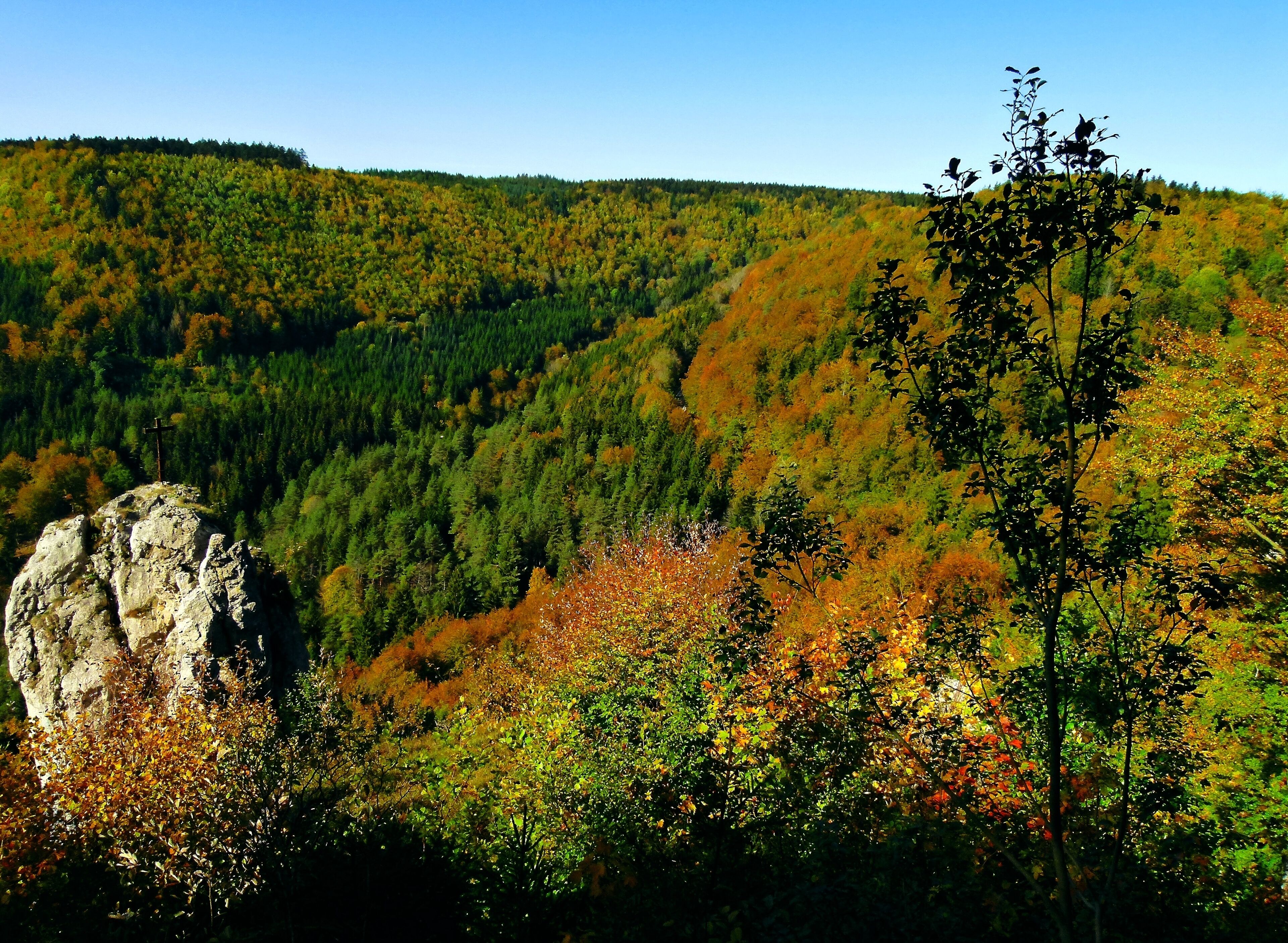 A mountainous landscape with autumn foliage, a prominent rock formation, and a cross on top.