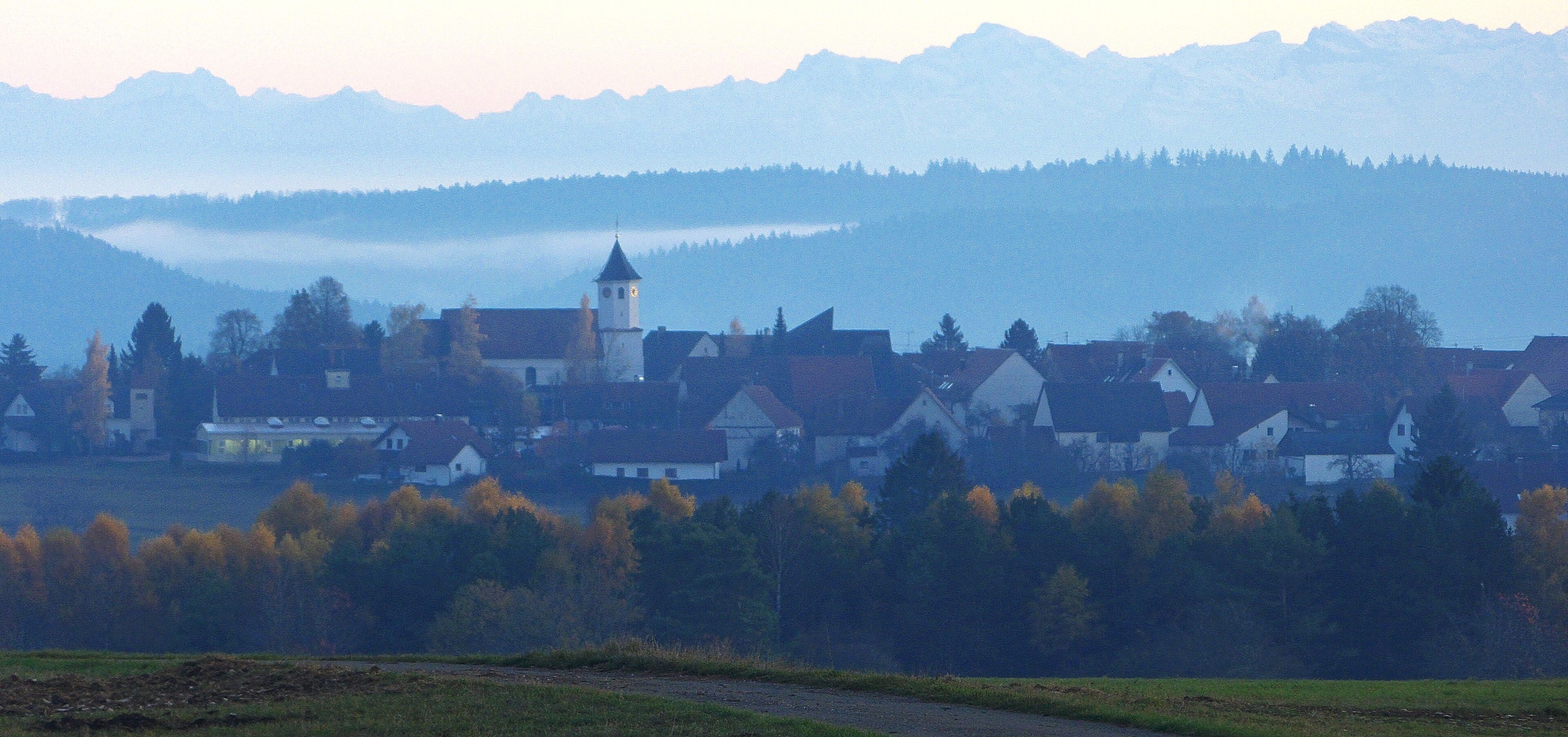 A village with a church tower, surrounded by autumn-colored trees and mountains in the background.