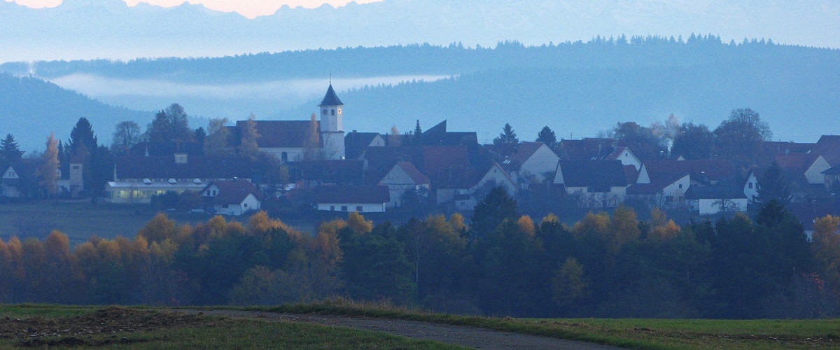 A village with a church tower, surrounded by autumn-colored trees and mountains in the background.