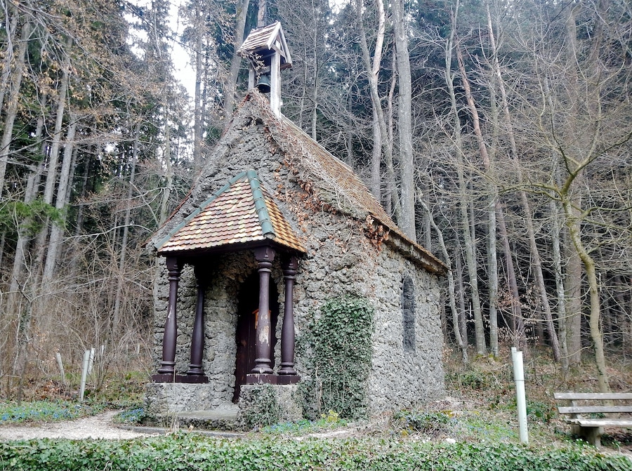 A small stone chapel with a tiled roof, surrounded by a forest.