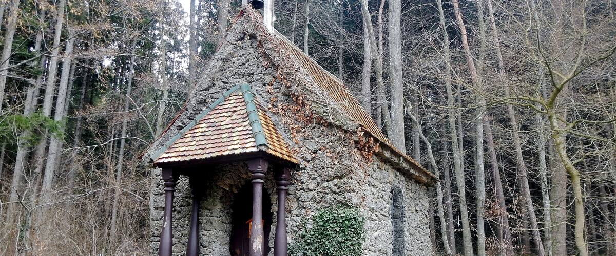 A small stone chapel with a tiled roof, surrounded by a forest.
