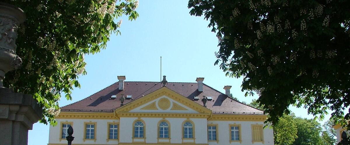 A large white building with yellow trim and a symmetrical facade, surrounded by a well-manicured lawn and trees.