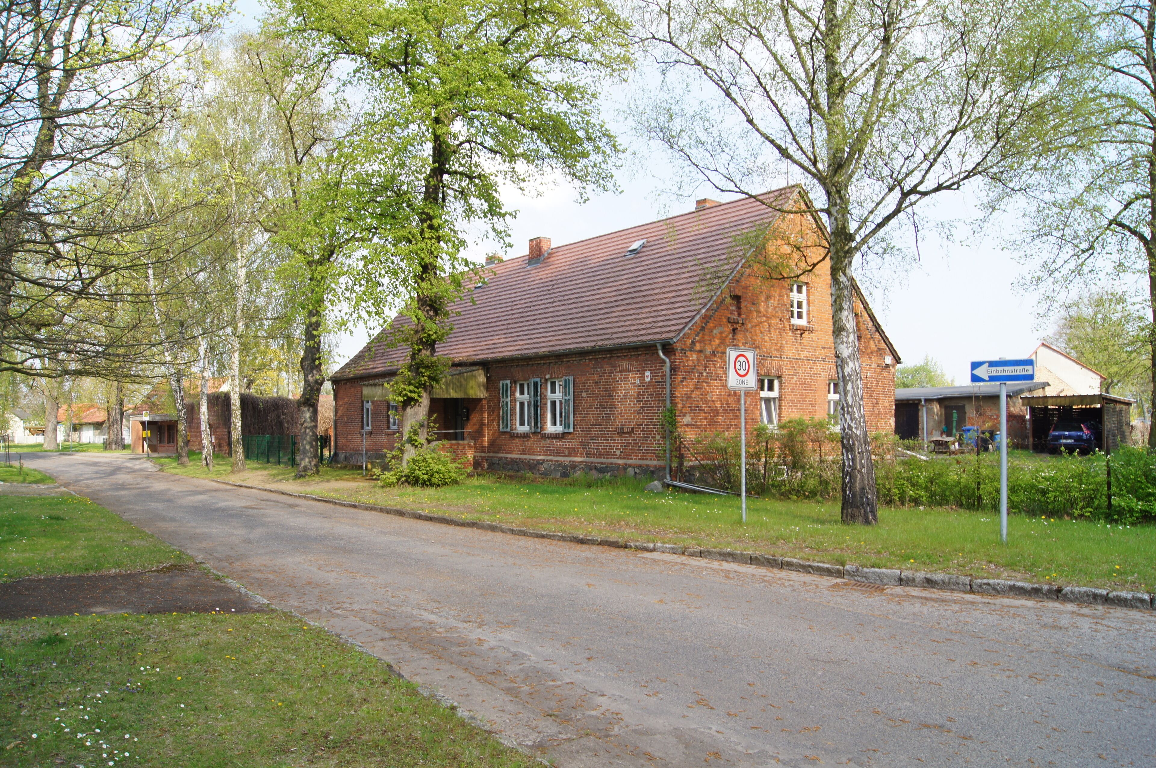 A quiet residential street with a brick house, a speed limit sign, and a directional sign.