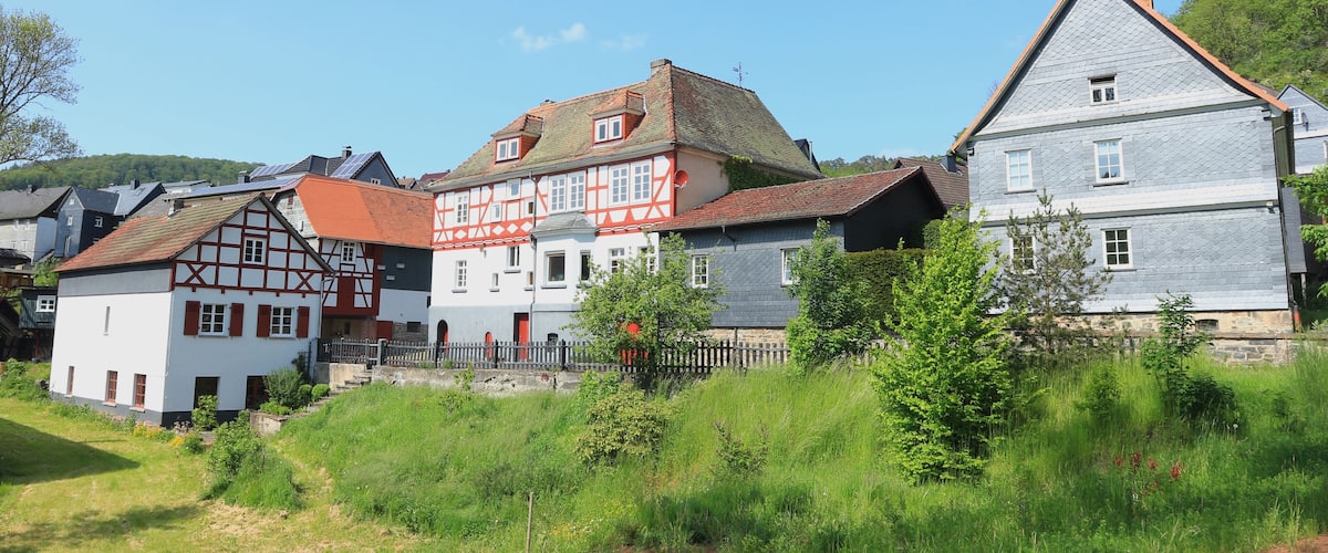 A row of traditional half-timbered houses with red and white facades, surrounded by lush greenery and a clear blue sky.