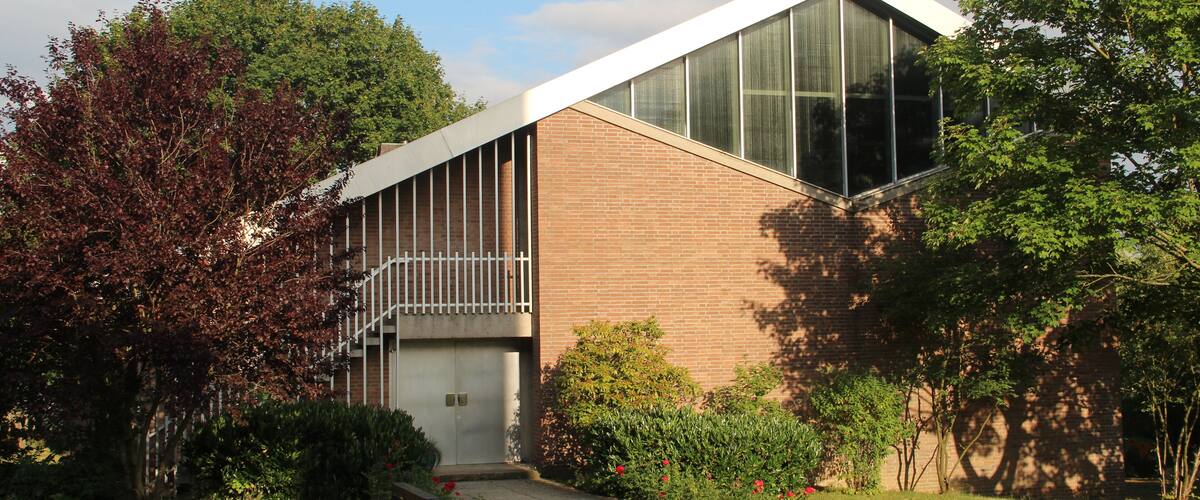 A modern building with large windows and a sloped roof, surrounded by trees and a grassy area.