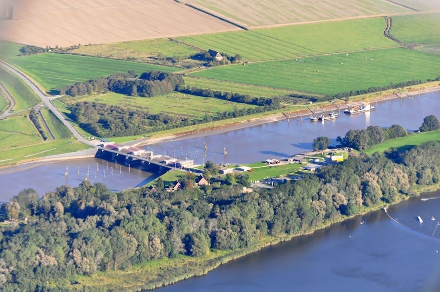 Vista aérea de un río con un puente, campos verdes y un pueblo pequeño con edificios y barcos.