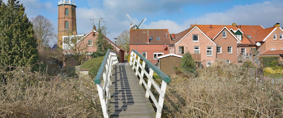 A wooden bridge with white railings leading to a town with brick buildings and a windmill.