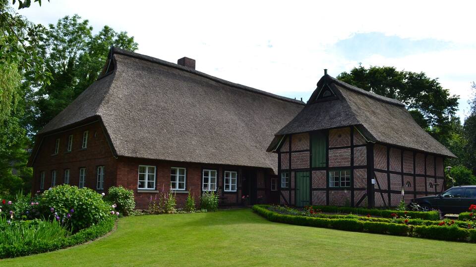 A traditional half-timbered house with a thatched roof, surrounded by a well-maintained garden and a lawn.