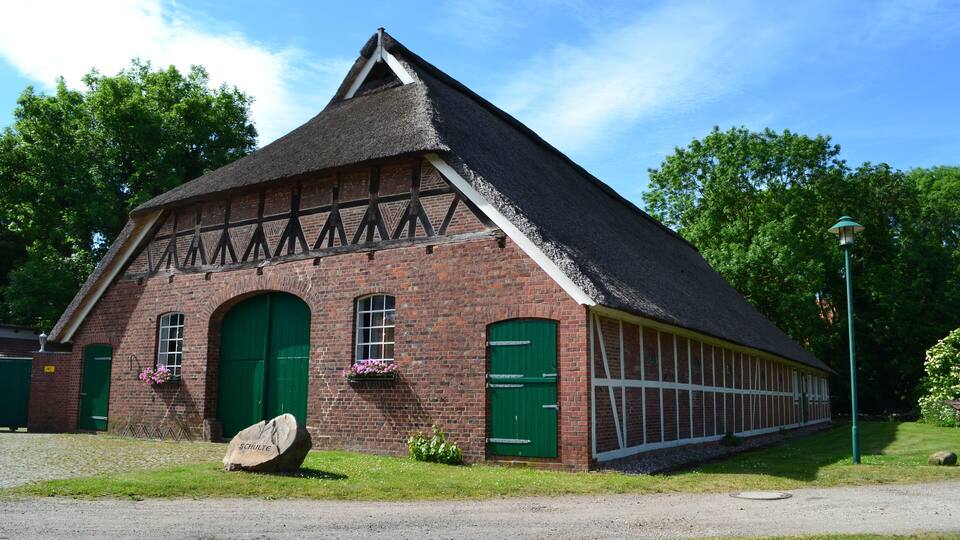 A traditional half-timbered barn with green doors and windows, surrounded by lush greenery.