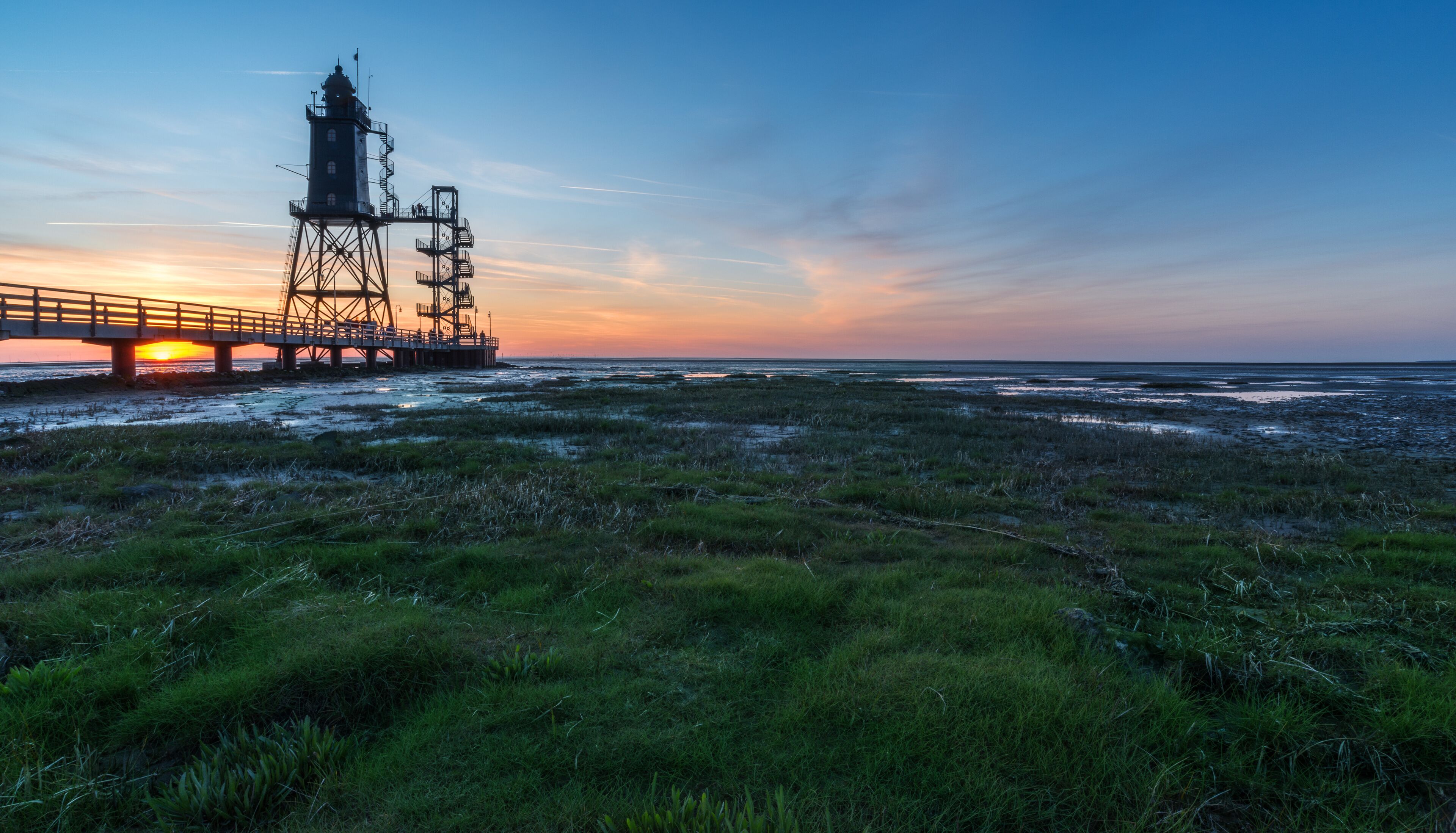 A coastal landscape with a pier and a lighthouse at sunset.