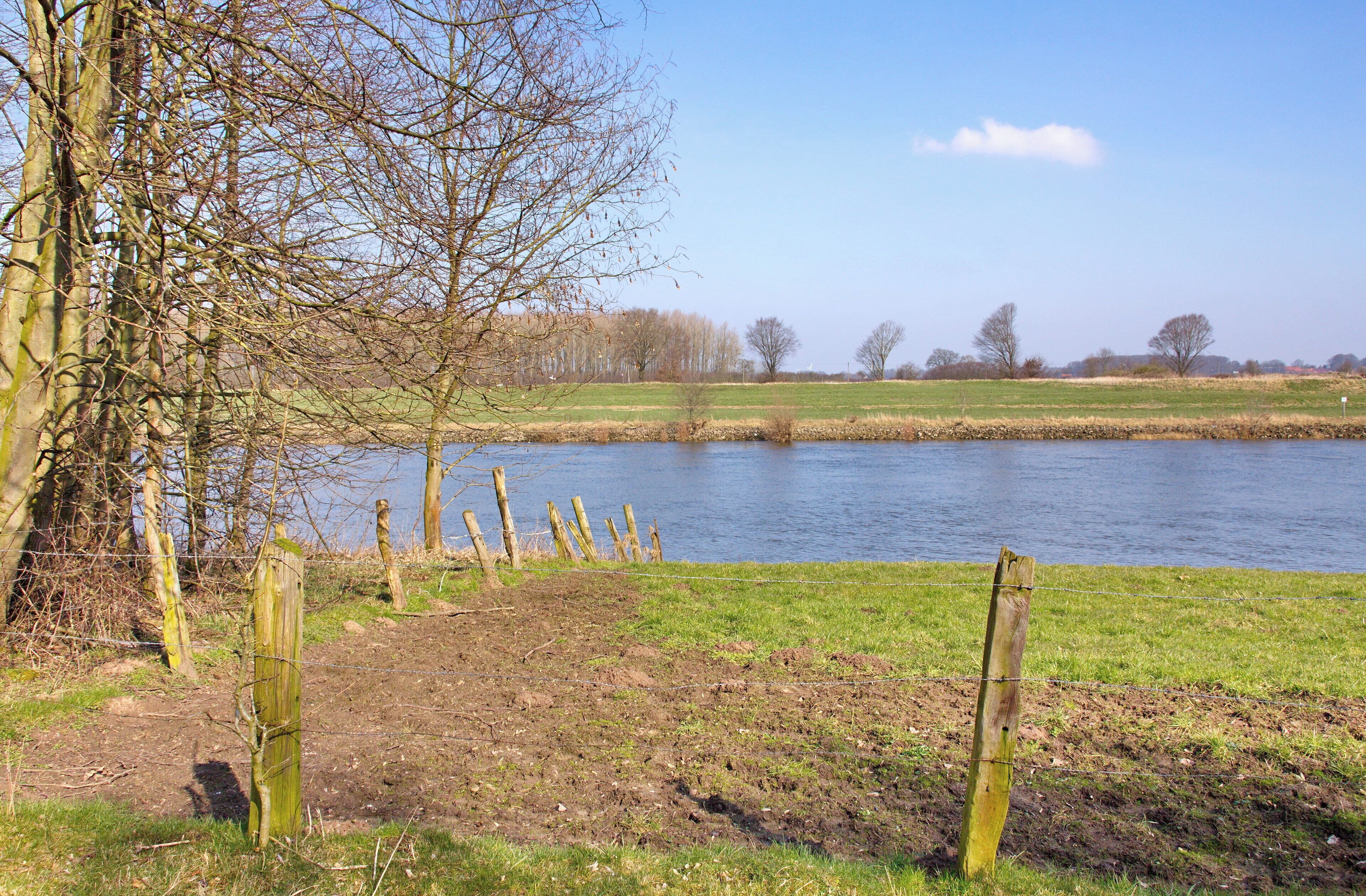 A river with a grassy bank, a wooden fence, and leafless trees in the background.