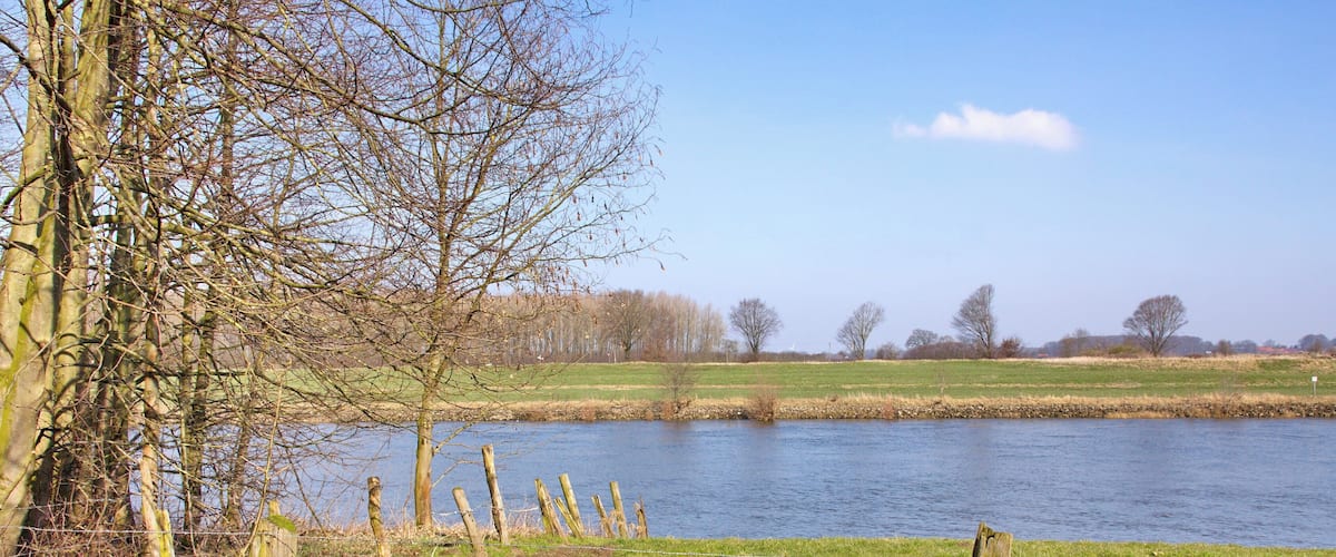 A river with a grassy bank, a wooden fence, and leafless trees in the background.