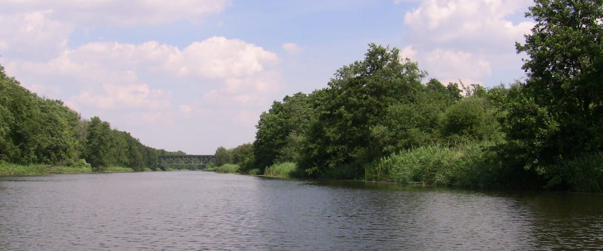 A calm river with dense greenery on both sides and a bridge in the distance.
