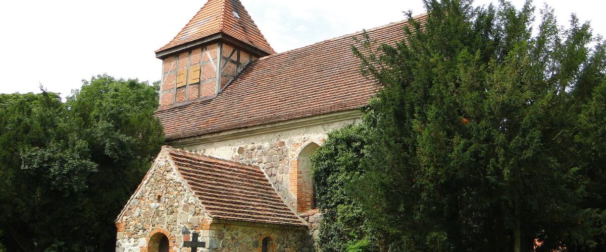 A church with a red-tiled roof and stone walls, surrounded by greenery and tombstones.