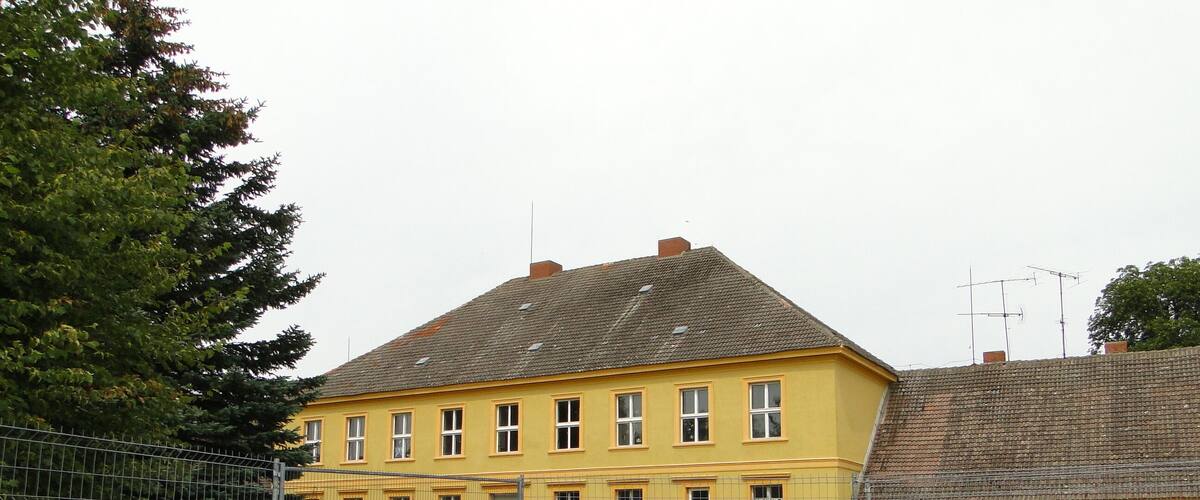 A yellow building with a grey roof, surrounded by a metal fence and greenery.