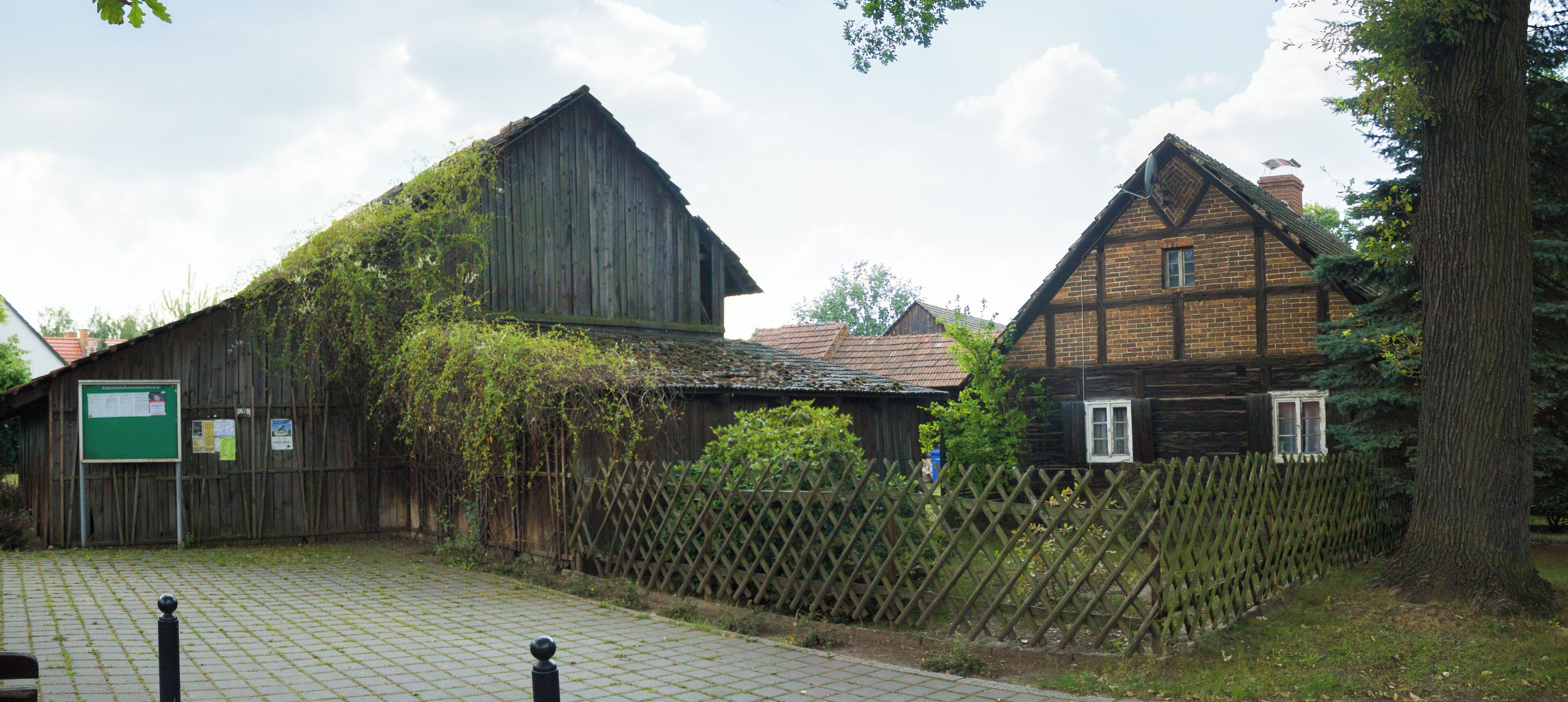 A rustic barn and a traditional house surrounded by a wooden fence and greenery.