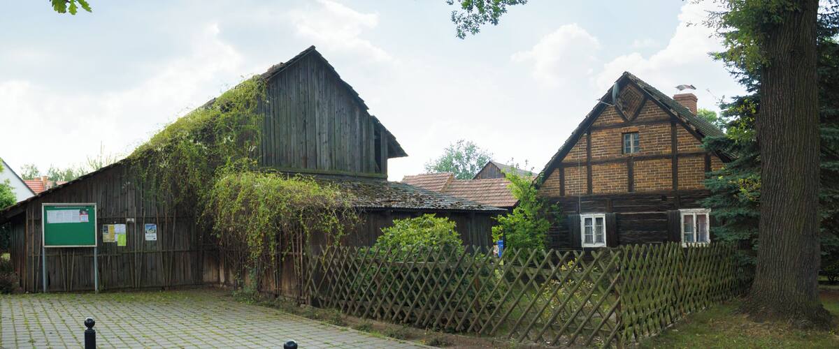 A rustic barn and a traditional house surrounded by a wooden fence and greenery.