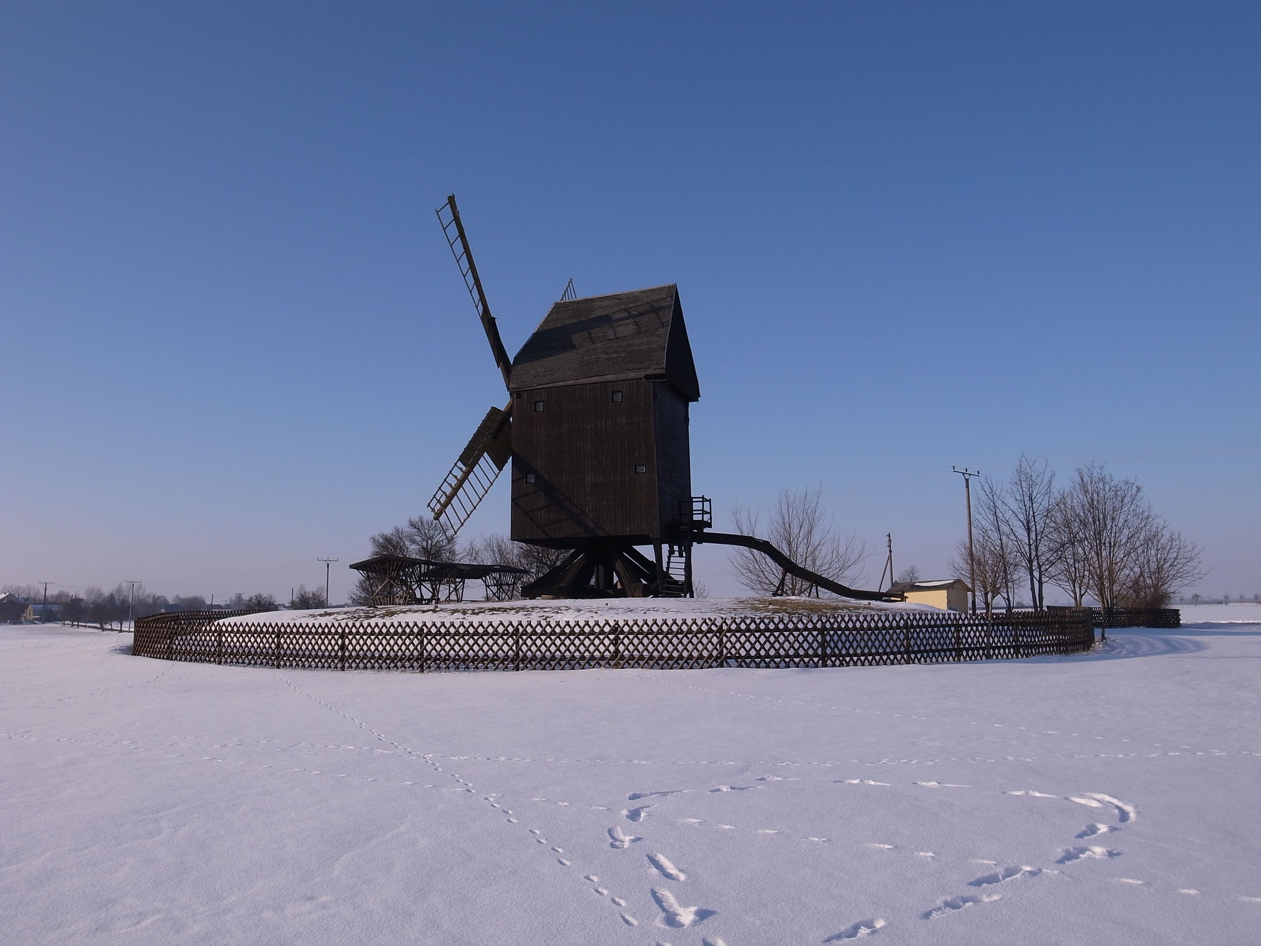 A wooden windmill in a snowy field with a fence around it.