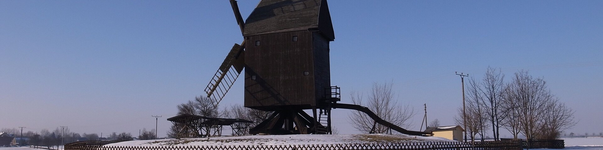 A wooden windmill in a snowy field with a fence around it.