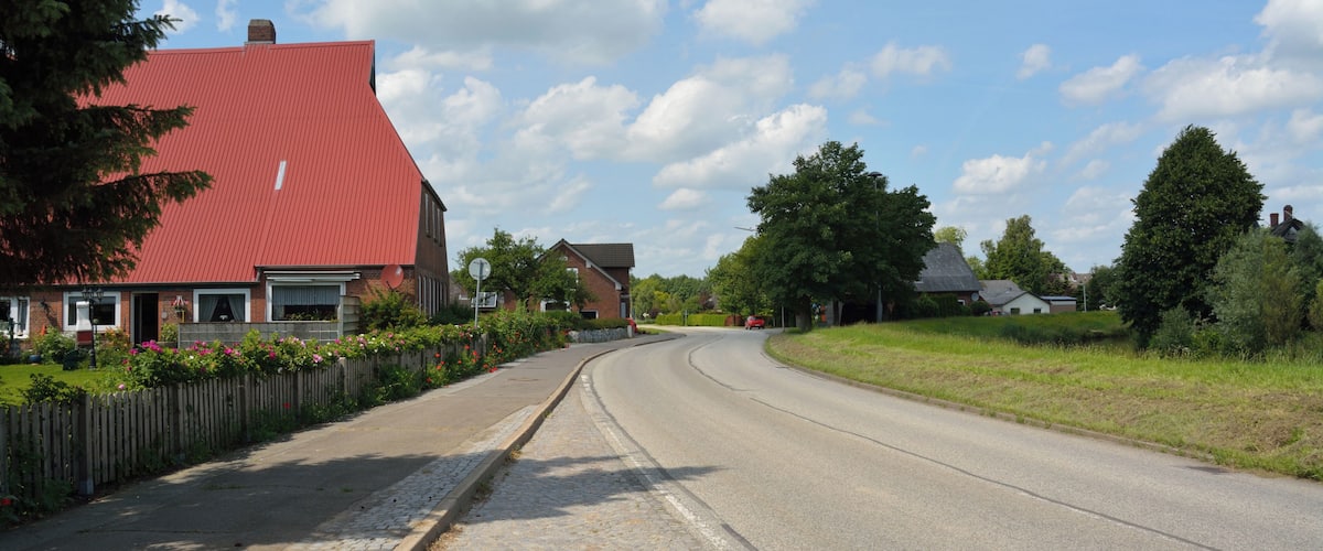 A rural road with houses, a cobblestone sidewalk, and a clear blue sky.