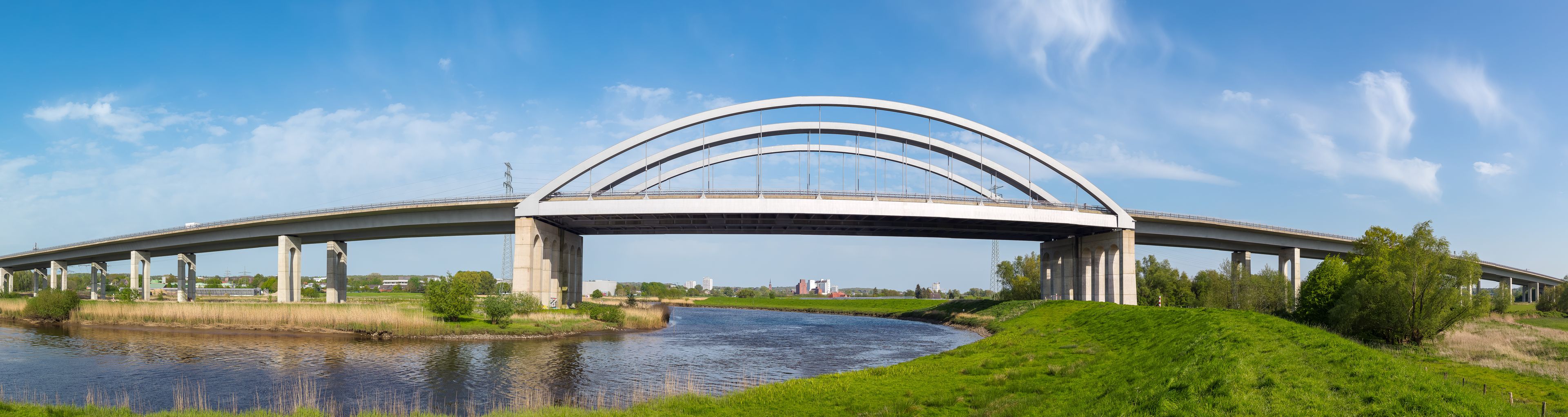 Un puente moderno con múltiples arcos sobre un río, rodeado de campos verdes y árboles.