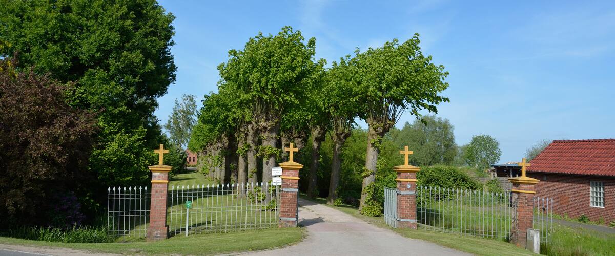A cemetery gate with a brick fence and crosses, surrounded by trees and a red-roofed building.