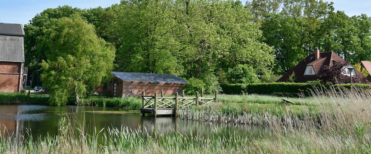 A river with a wooden bridge, a brick building, and a house with a red roof.
