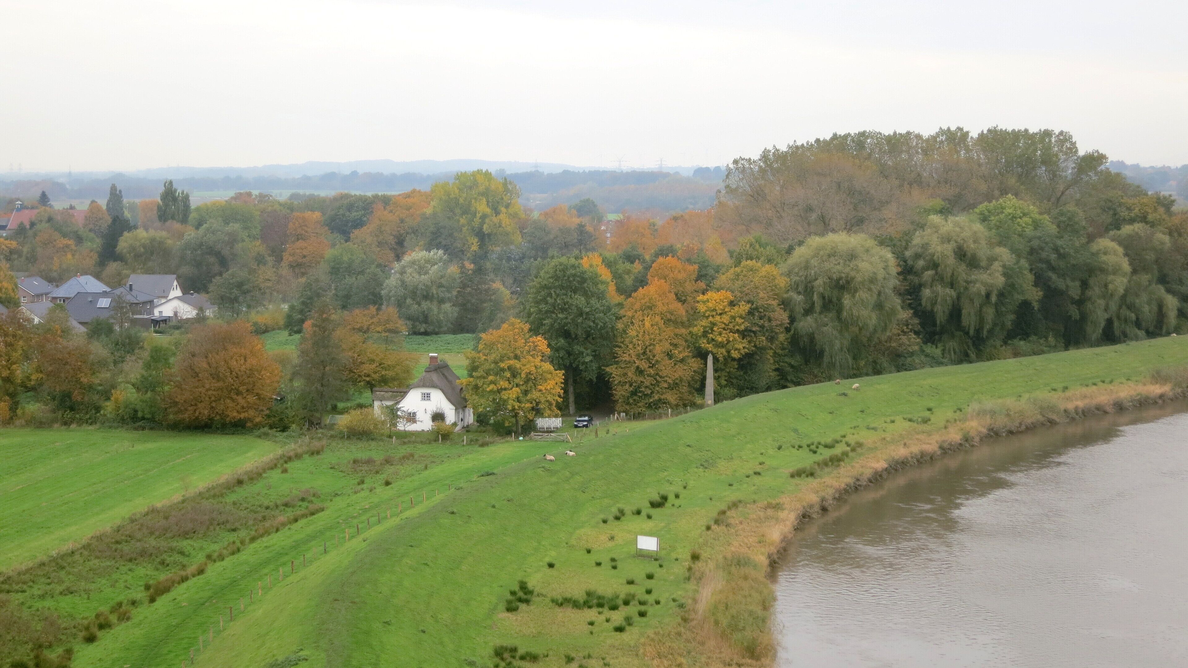A rural landscape with a river, rolling green fields, and autumn-colored trees.