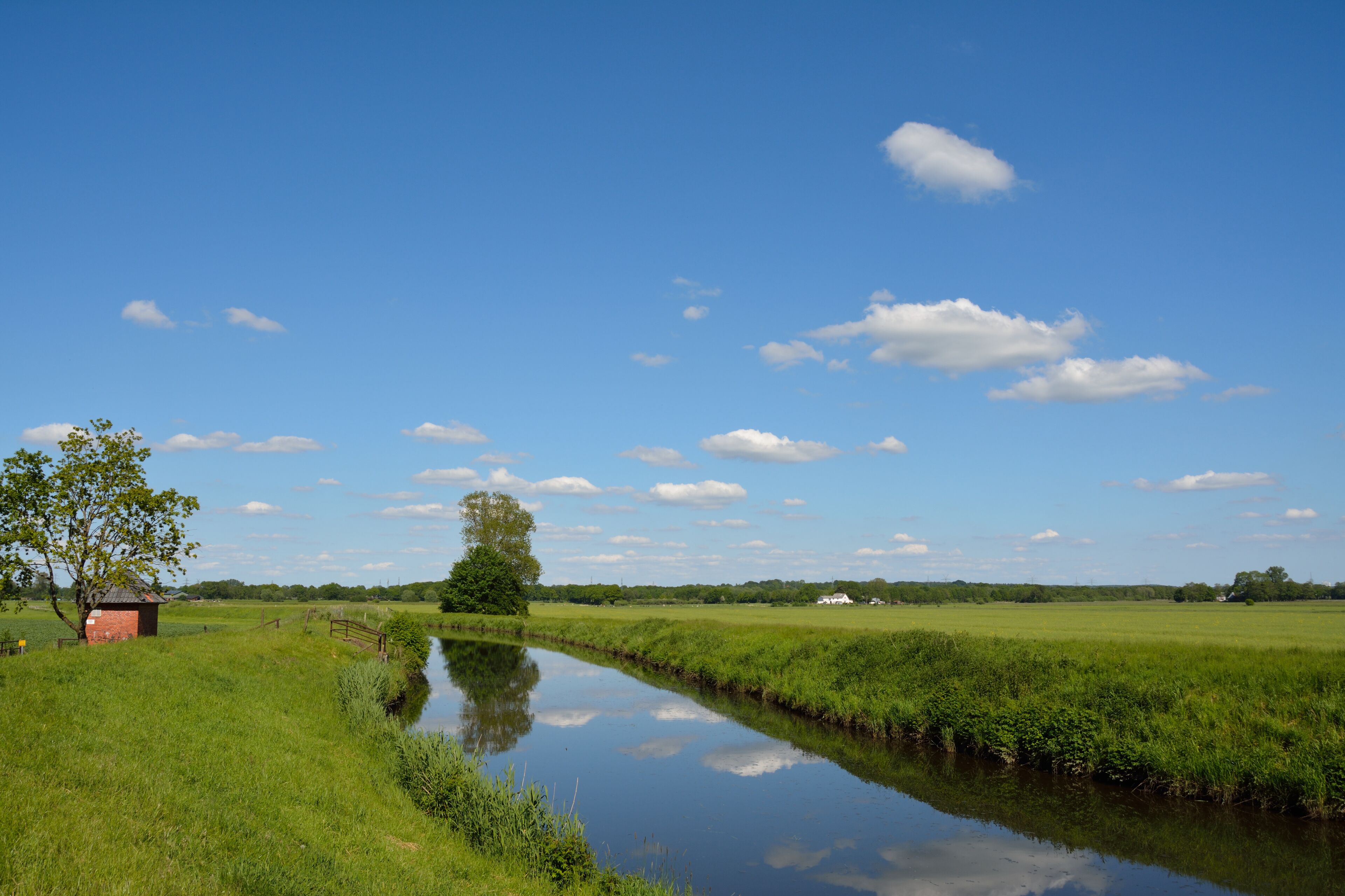 A calm river, green fields, a small red building, and a clear blue sky with clouds.