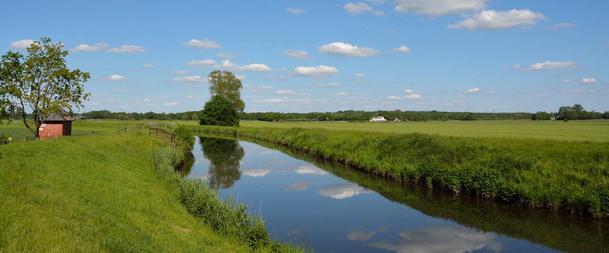 A calm river, green fields, a small red building, and a clear blue sky with clouds.