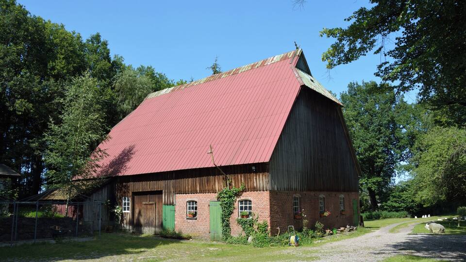 A red-roofed barn with green doors and windows, surrounded by trees and a gravel path.