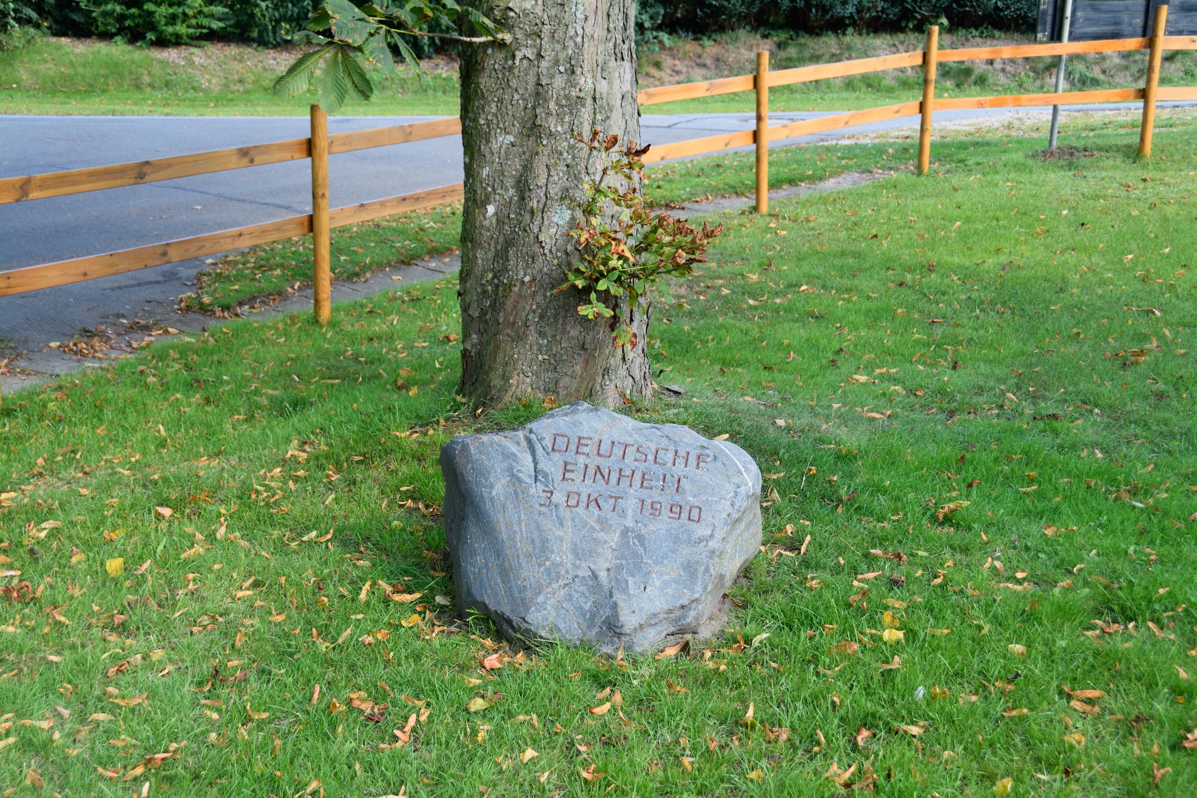 A stone with German text and a date, surrounded by grass and a tree.