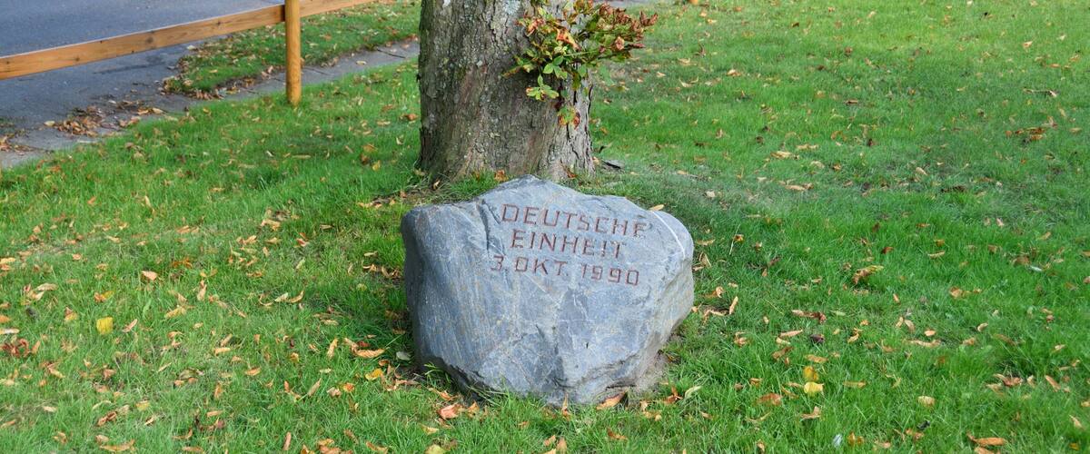 A stone with German text and a date, surrounded by grass and a tree.