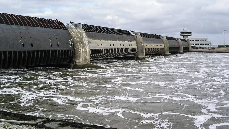 A large industrial structure with multiple cylindrical sections, situated by a body of water.