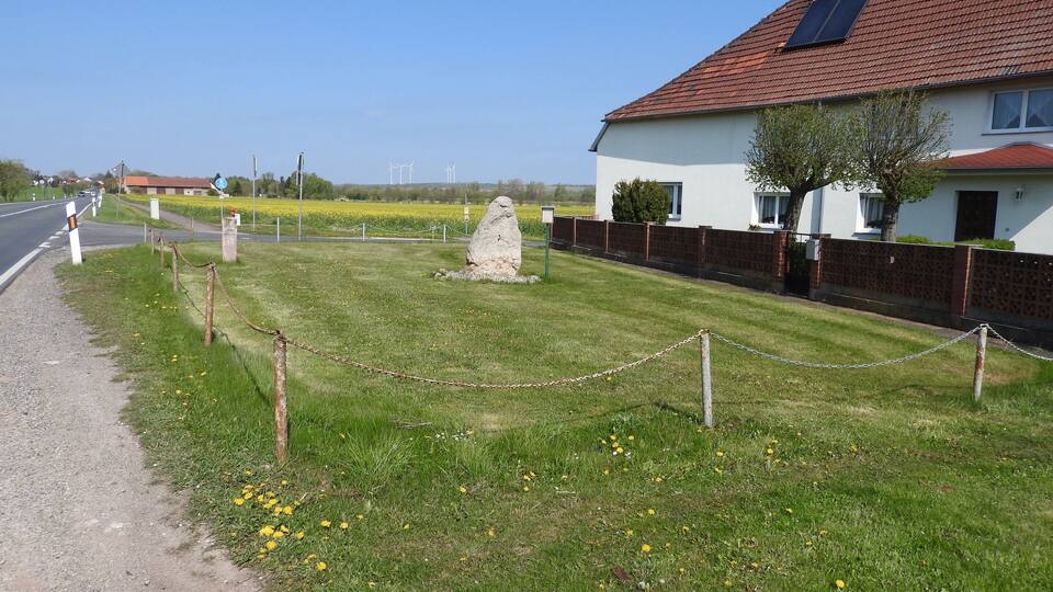 A rural landscape with a white house, a stone monument, and a field of yellow flowers.