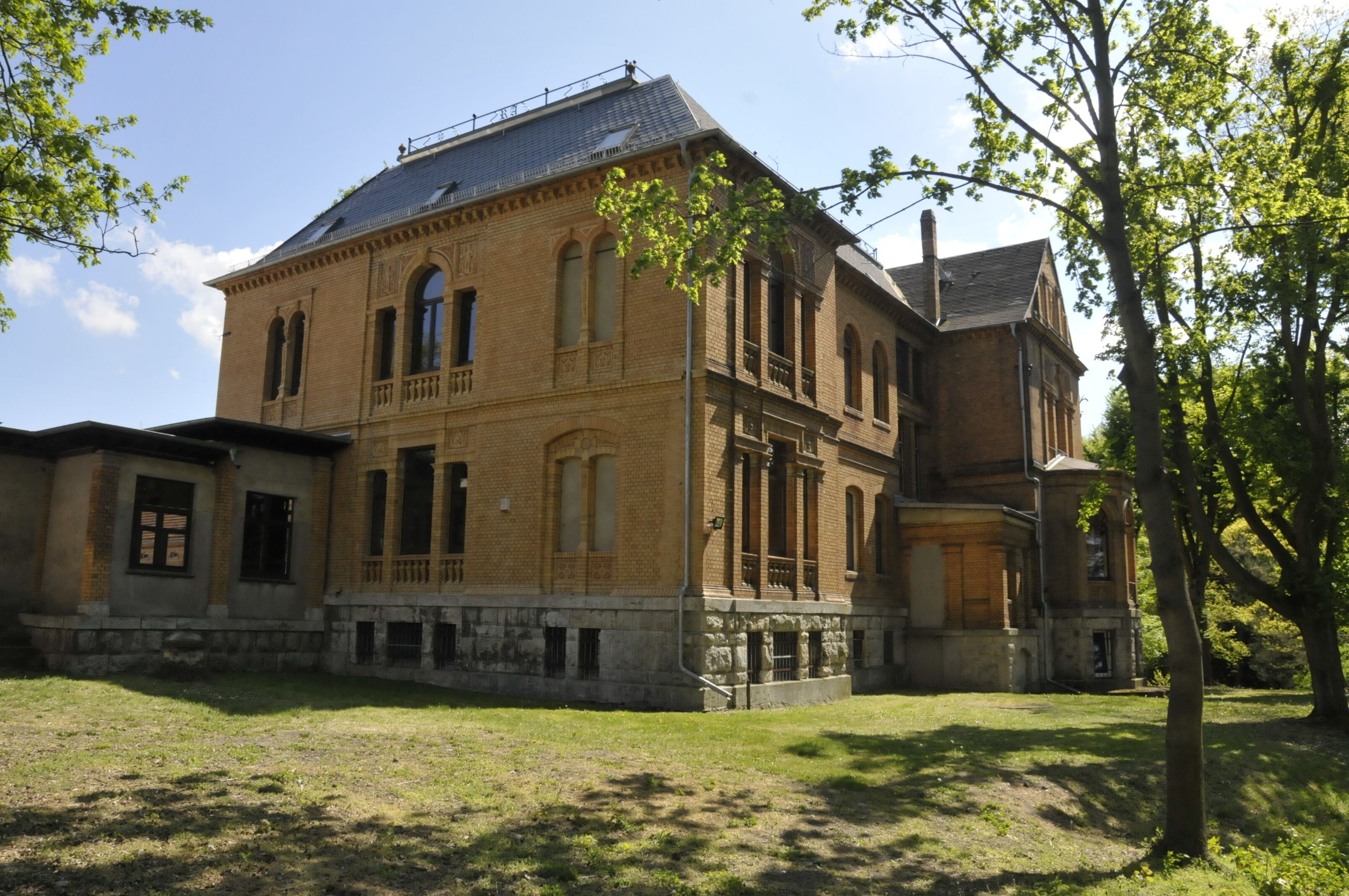 A historic brick building with arched windows and a balcony, surrounded by a grassy area and trees.