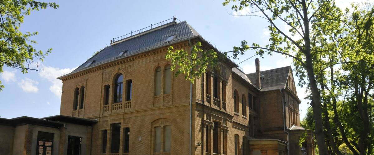 A historic brick building with arched windows and a balcony, surrounded by a grassy area and trees.