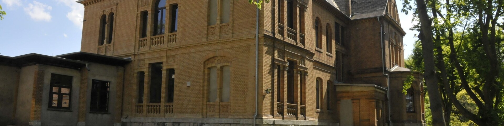 A historic brick building with arched windows and a balcony, surrounded by a grassy area and trees.