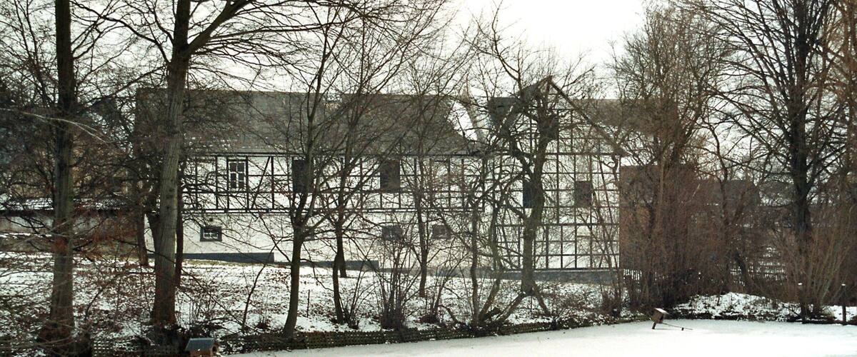 A snow-covered yard with a wooden fence, leafless trees, and a building in the background.