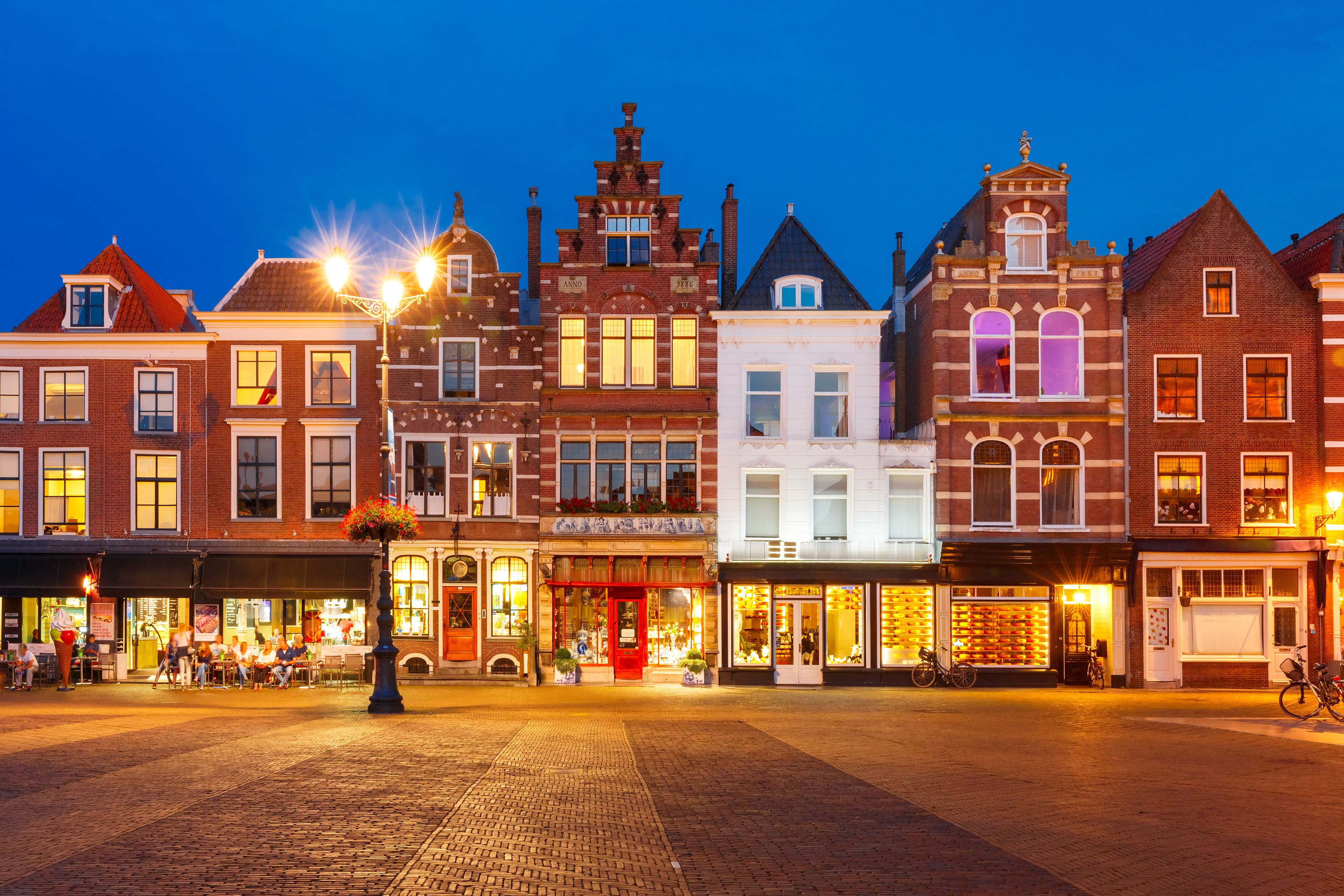 A historic European town square at dusk with traditional brick buildings, illuminated windows, and a cobblestone pavement.