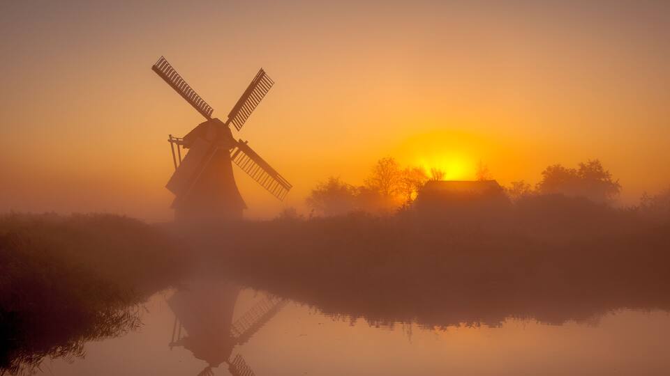 A windmill silhouetted against a sunrise, with a calm body of water reflecting its image.