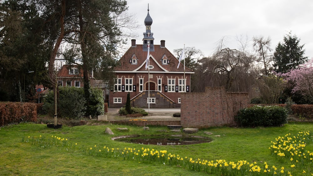 A large brick house with a tower, surrounded by a well-maintained garden with daffodils and a pond.