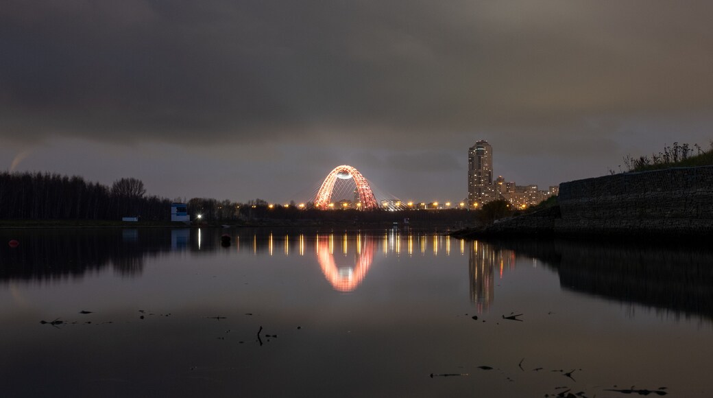 A calm lake reflecting a brightly lit Ferris wheel and a tall building.
