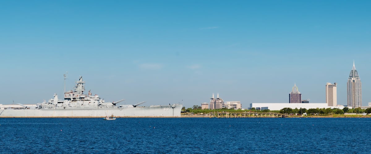 A large naval ship docked at a port with a city skyline in the background.